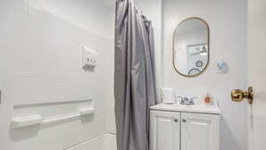 Un baño con paredes de azulejos blancos, una bañera, una cortina de ducha, un lavamanos con espejo y un perchero para toallas.