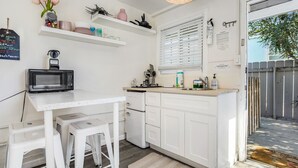 Una cocina compacta con armarios blancos, una pequeña encimera y un microondas sobre una mesa. Hay taburetes, un lavamanos y una ventana con persianas.