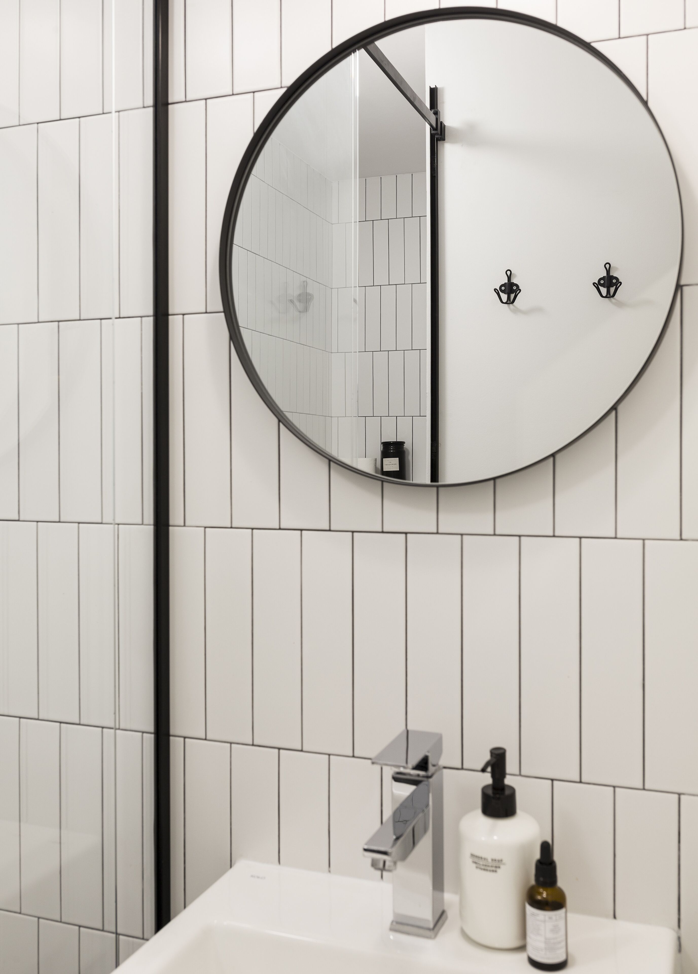 Een badkamer met een ronde spiegel, witte tegels, een moderne kraan en twee flessen toiletartikelen.