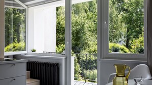 Et moderne værelse med et stort vindue der giver udsigt til en frodig have. Der er en radiator, et lille bord med en kande samt en stol.