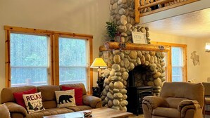 A spacious living room with a stone fireplace, a high ceiling, and a wooden staircase.