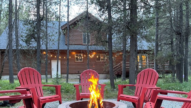 A log cabin with a stone fire pit surrounded by red Adirondack chairs.