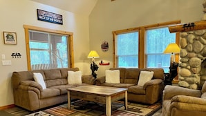 A cozy living room with two brown sofas, a wooden coffee table, and a patterned rug. There are two windows with blinds, a stone wall, and a welcome sign.