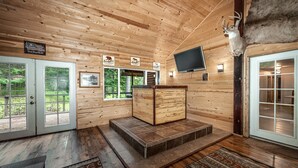 Interior de una cabaña de troncos con chimenea de piedra, paredes de madera, una cabeza de venado montada y un televisor.