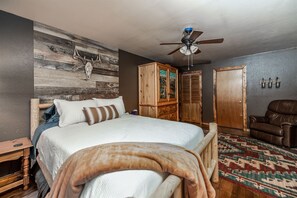 Ein Schlafzimmer mit einem Bett, einem Holzkopfende, einem Deckenventilator, einem Kleiderschrank, einem braunen Ledersessel und einem gemusterten Teppich.