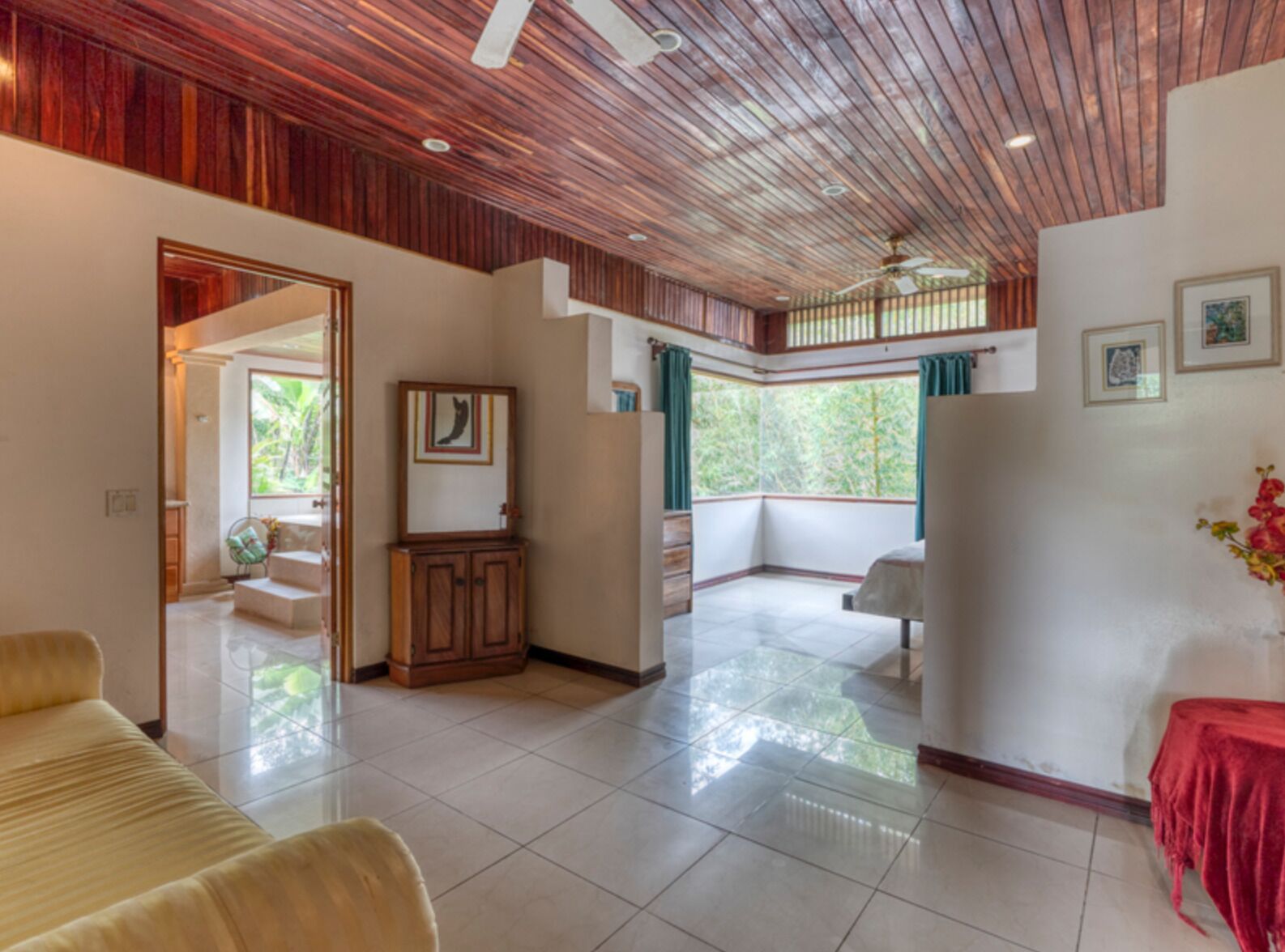 A spacious room with a wooden ceiling, a balcony with a view of greenery, and a red table.
