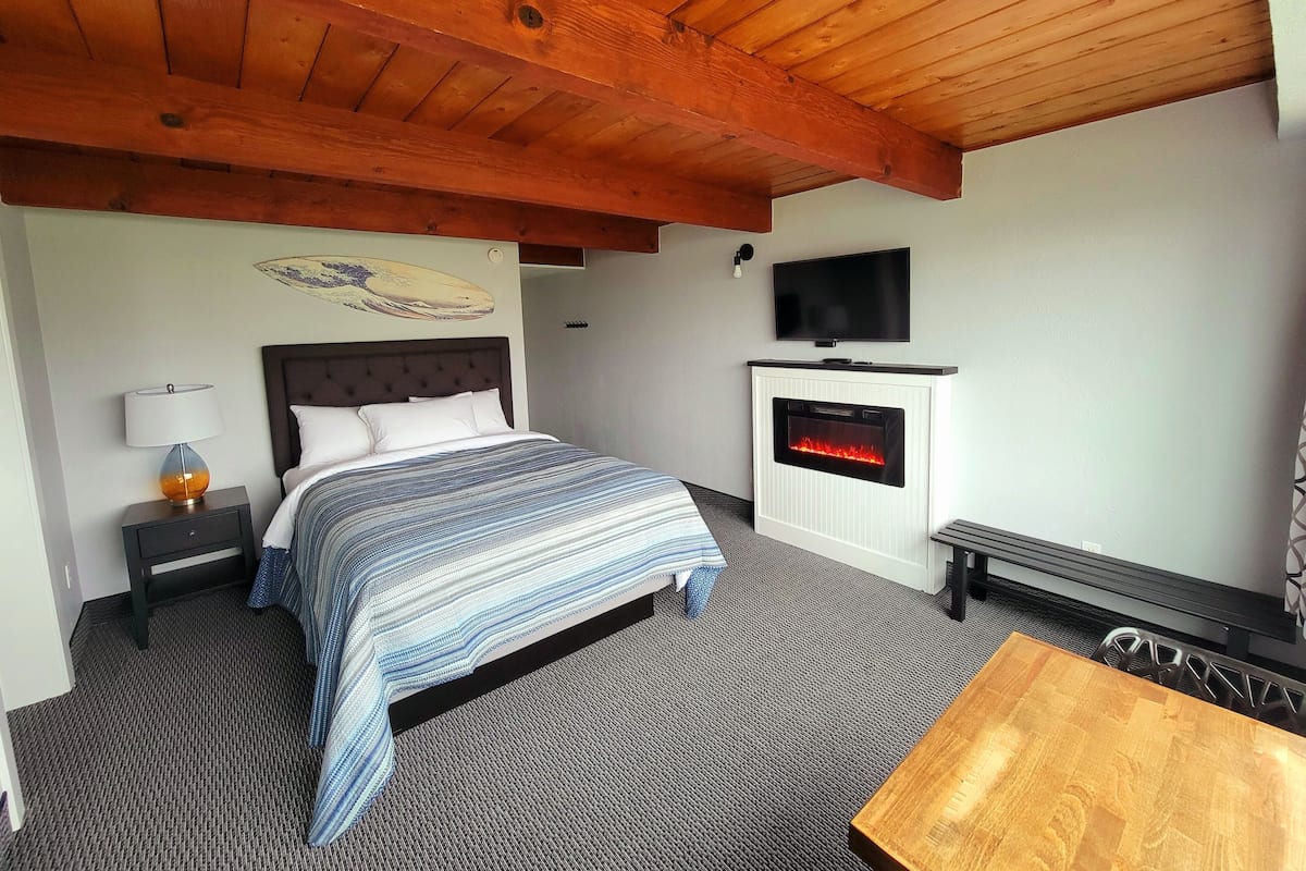 A bedroom with a bed, a TV mounted above a fireplace, a nightstand with a lamp, and a wooden ceiling.