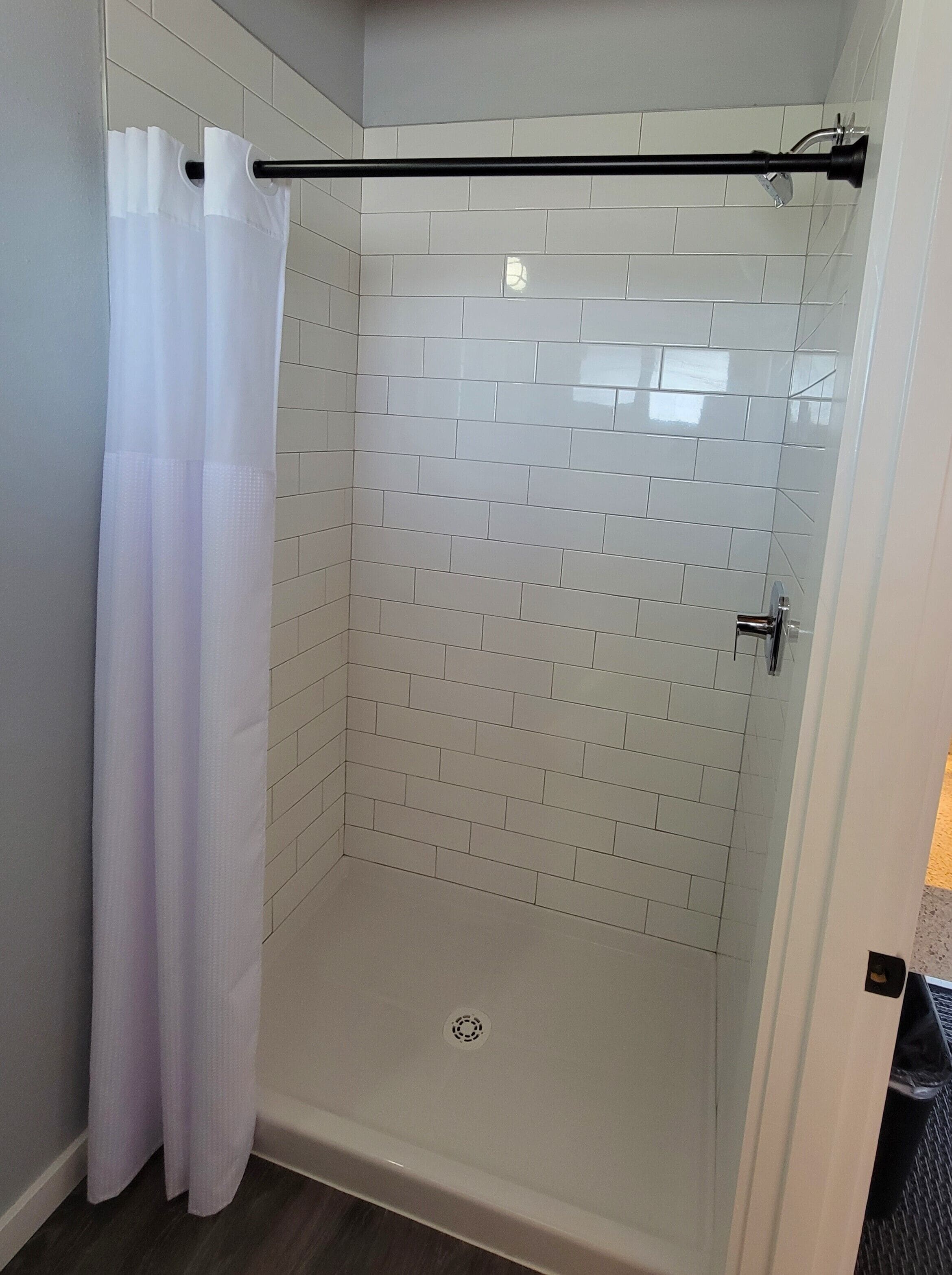 Une douche fermée par des parois de verre, avec des murs recouverts de carreaux blancs et un rideau de douche blanc.