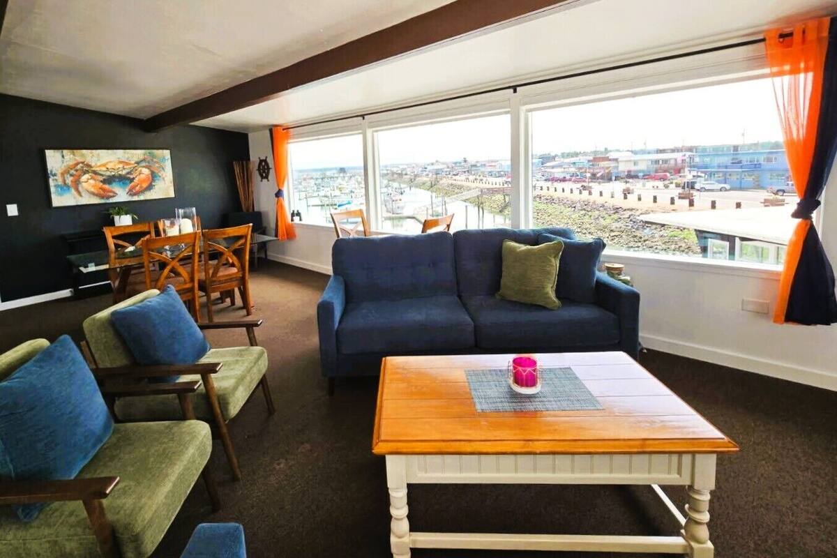 A living room with a blue sofa, wooden chairs, a wooden coffee table, and a large window with a view of a cityscape.
