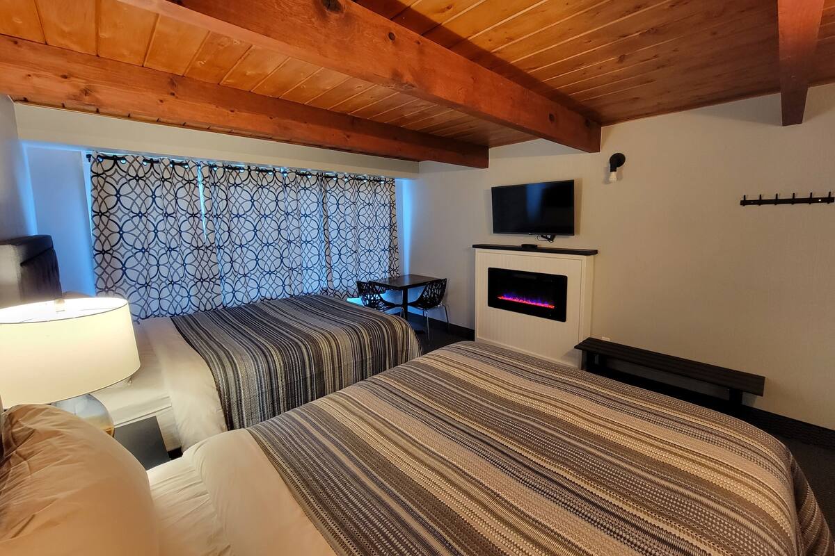 A hotel room with a wooden ceiling, a bed with striped bedding, a TV mounted above a fireplace, and a patterned curtain.