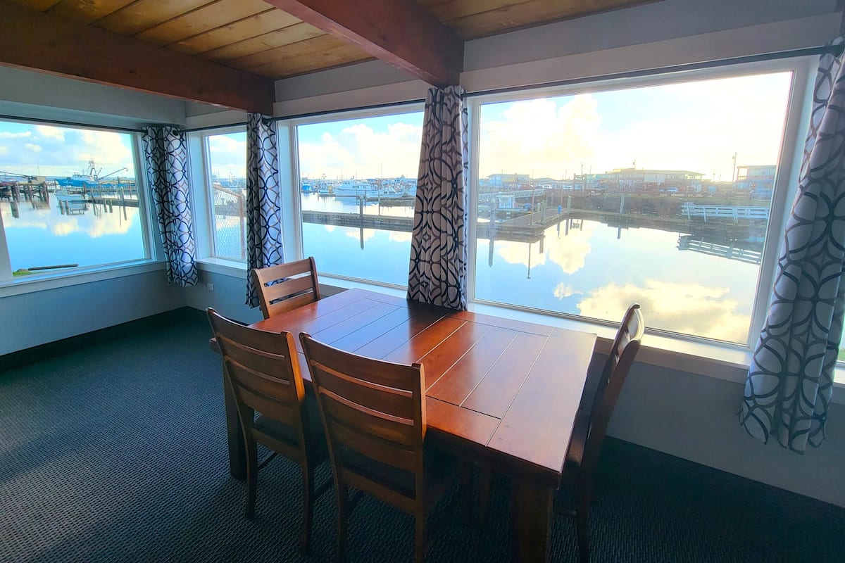 A dining area with a wooden table and chairs, large windows with patterned curtains, and a view of a marina with boats.