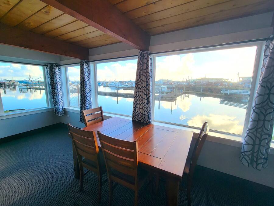 Un comedor con una mesa y sillas de madera, ventanas amplias con cortinas estampadas y vista a un puerto deportivo con barcos.