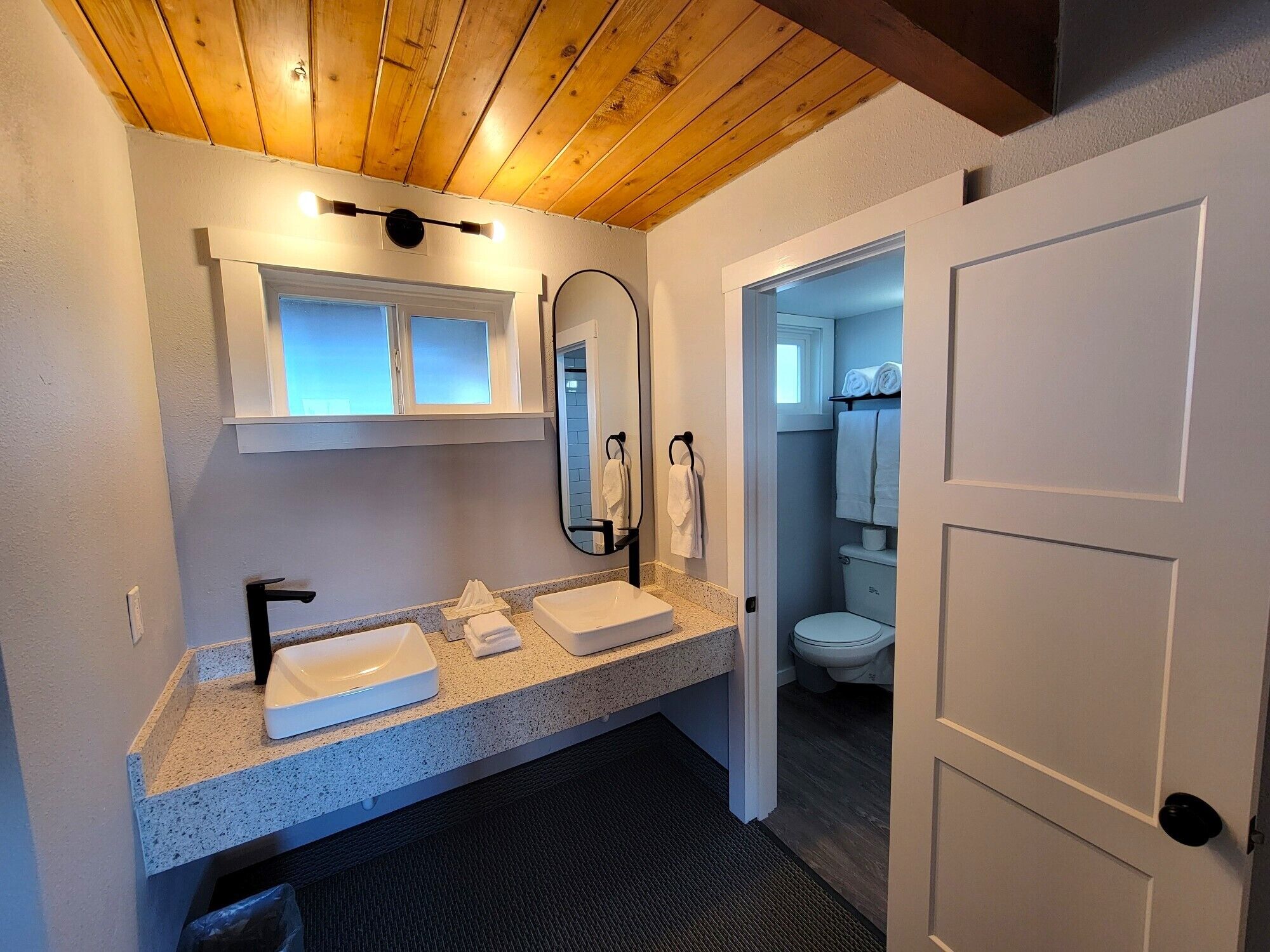 A modern bathroom with a wooden ceiling, a double sink vanity, a mirror, and a toilet visible through an open door.