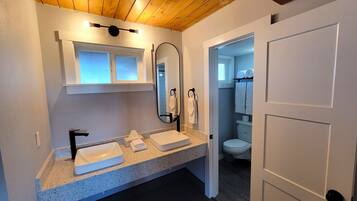 Une salle de bain moderne avec un plafond en bois, un meuble-lavabo à deux vasques, un miroir et des toilettes visibles à travers une porte ouverte.