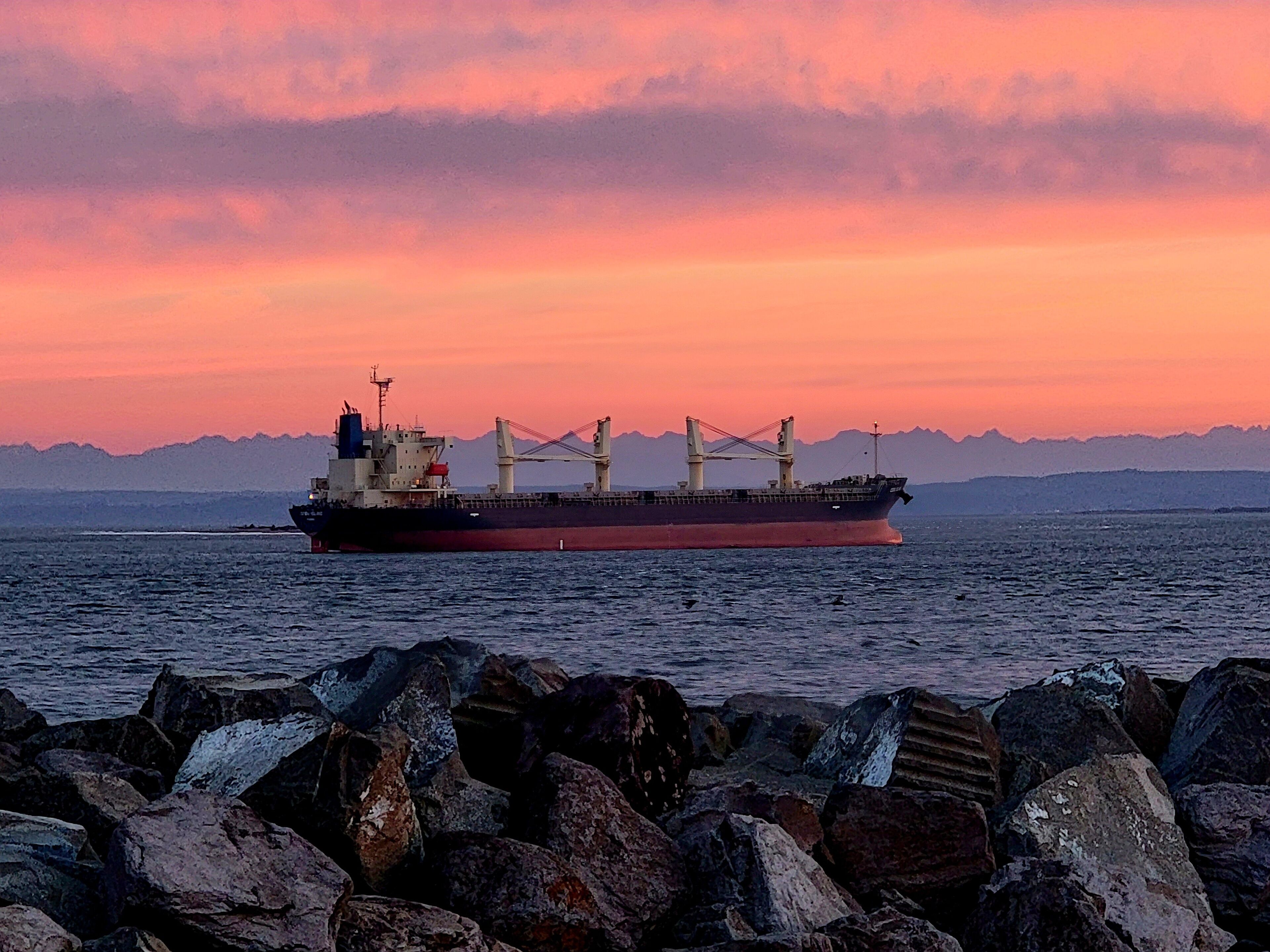 Een vrachtschip op zee tijdens de zonsondergang, met rotsachtige oevers op de voorgrond.