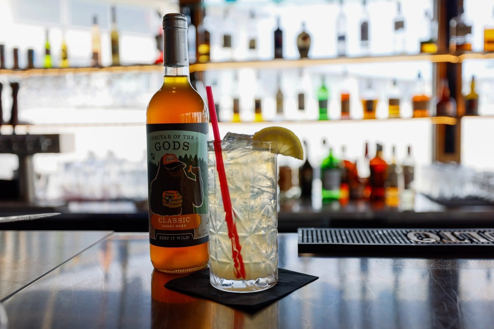 A bottle of Nectar of the Gods Classic Honey Mead and a glass with a red straw and lemon slice on a bar counter.