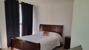 A neatly made bed with a wooden headboard, a bedside table, and a window with dark curtains. - Tierra Adentro Bed & Breakfast (Naguabo)