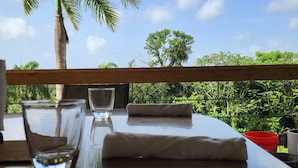 A table with a glass of water, a folded towel, and a red bowl, set against a backdrop of a palm tree and lush greenery. - Tierra Adentro Bed & Breakfast (Naguabo)