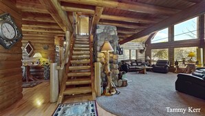 A log cabin interior with a wooden staircase, stone wall, and a cozy living area with a fireplace.