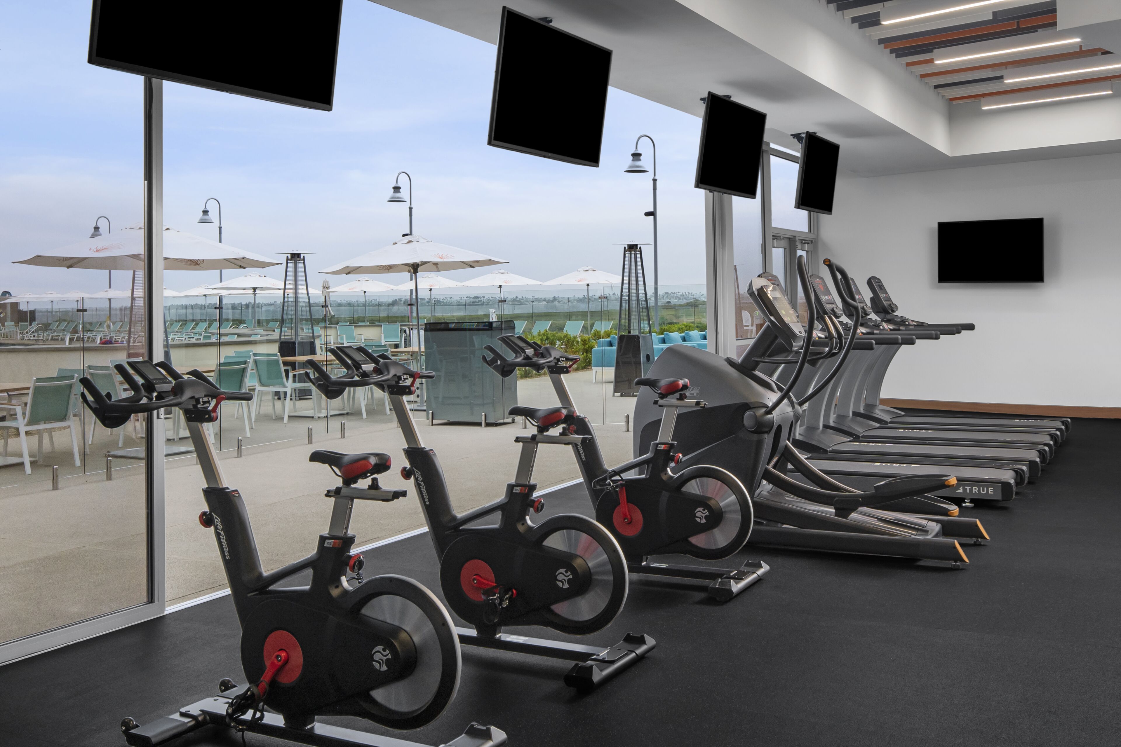 Un gimnasio con bicicletas estáticas y cintas de correr, ventanas amplias y zona de estar exterior con sombrillas.