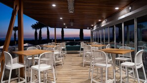 Un espace de restauration en plein air avec des tables en bois et des chaises blanches, offrant une vue panoramique sur des palmiers et un coucher de soleil.
