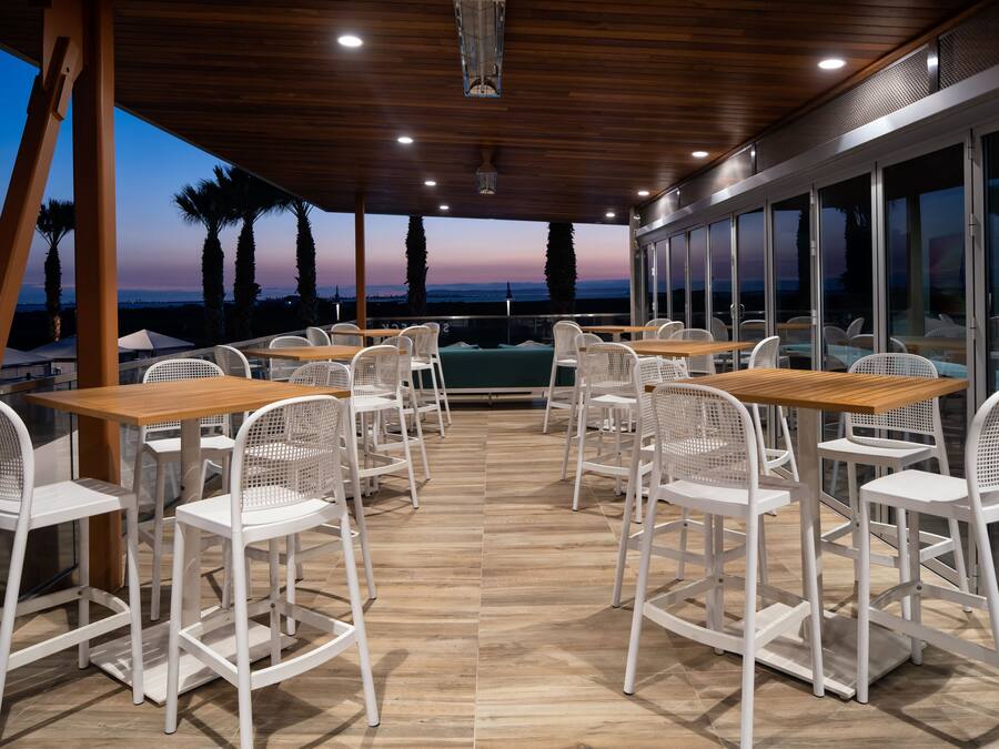 An outdoor dining area with wooden tables and white chairs, overlooking a scenic view with palm trees and a sunset.