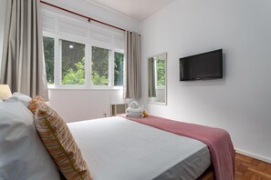 A hotel room with a bed, a TV mounted on the wall, a window with curtains, and a small table with towels.