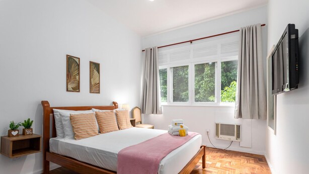 Un dormitorio con una cama de madera, una ventana con cortinas, un televisor montado en la pared y una mesita de noche con una planta.