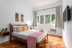 A bedroom with a wooden bed, a window with curtains, a wall-mounted TV, and a small bedside table with a plant.