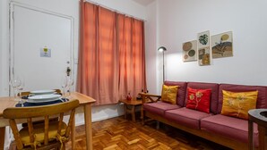 Sala de estar con una mesa de madera dispuesta para comer, un sofá con cojines, una lámpara de pie y obras de arte en la pared.