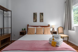 A neatly arranged hotel room with a bed, a side table with a lamp, a chair, a shelf with hangers, and a window with curtains.