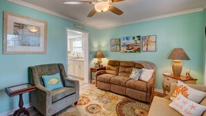 A living room with a ceiling fan, a sofa, a chair, and a coffee table. There are framed pictures on the wall and a rug on the floor. - Bungalow on 5th (Kure Beach)