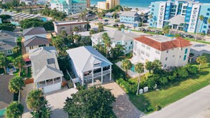 An aerial view of a coastal residential area with various buildings, including a prominent white house with a red roof and a large swimming pool, surrounded by palm trees and a clear view of the ocean.