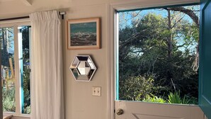 A room with a door, a window with a view of greenery, a hexagonal mirror, and a painting on the wall. - Quintessential Carmel Writer's Hideaway with Private Garden and Patio. (Carmel-by-the-Sea)