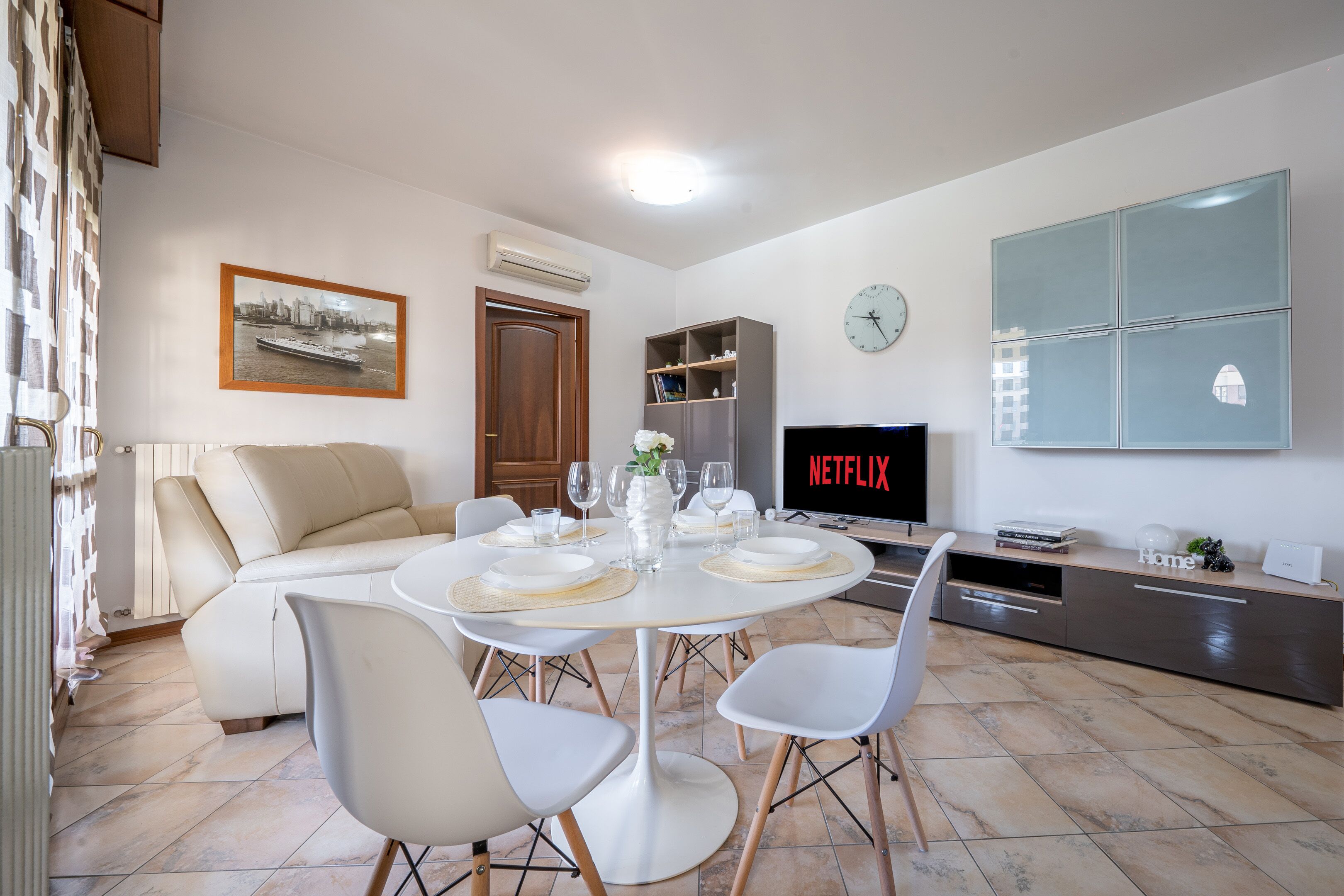 A modern living room with a dining table set for four, a sofa, a TV, and a cabinet with a clock.