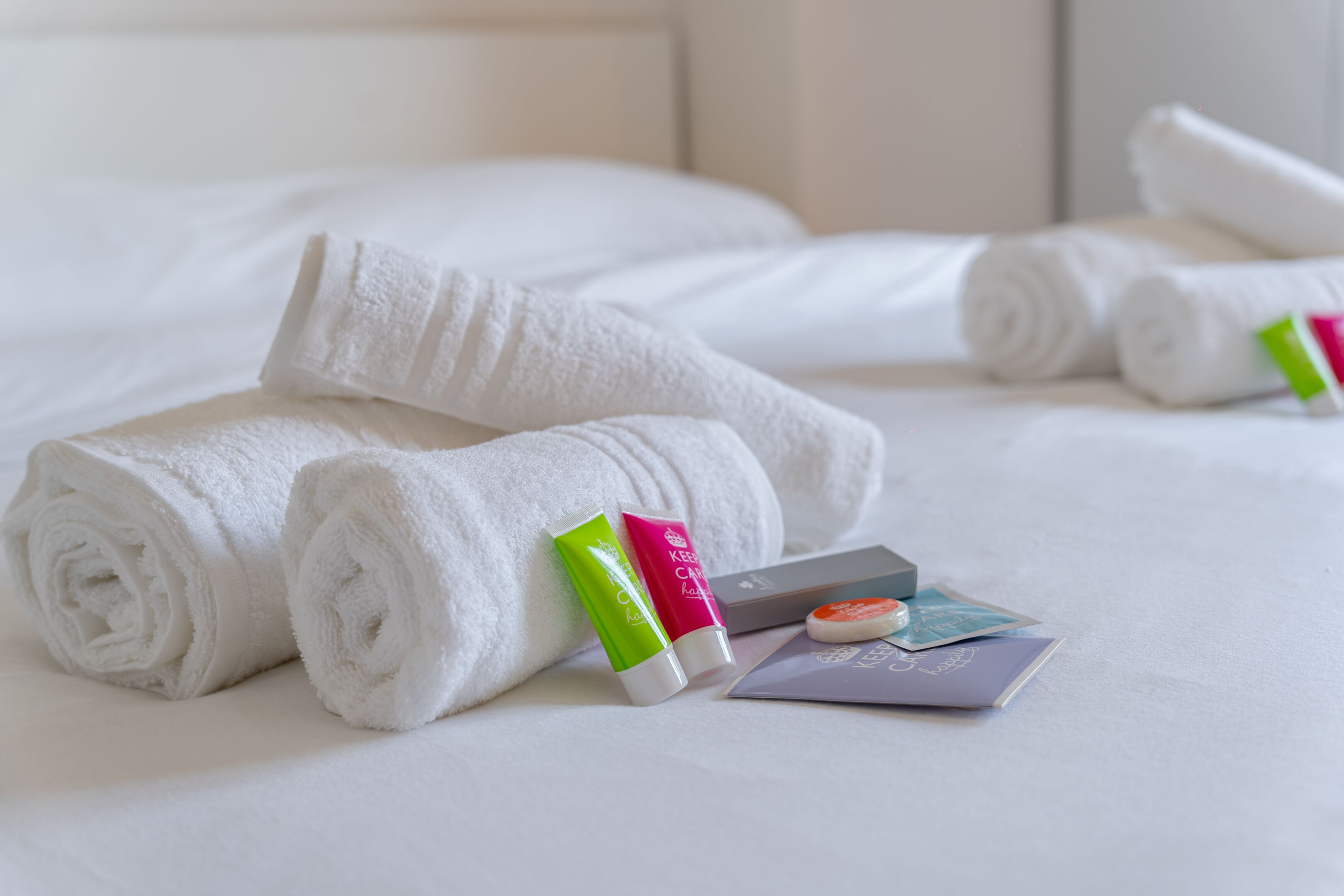 Neatly rolled white towels, a green and pink bottle, and a purple booklet on a bed.