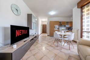 Ein modernes Wohnzimmer mit einem Flachbildfernseher, der 'NETFLIX' zeigt, einem Essbereich mit einem weißen Tisch und Stühlen sowie einer Küche im Hintergrund.