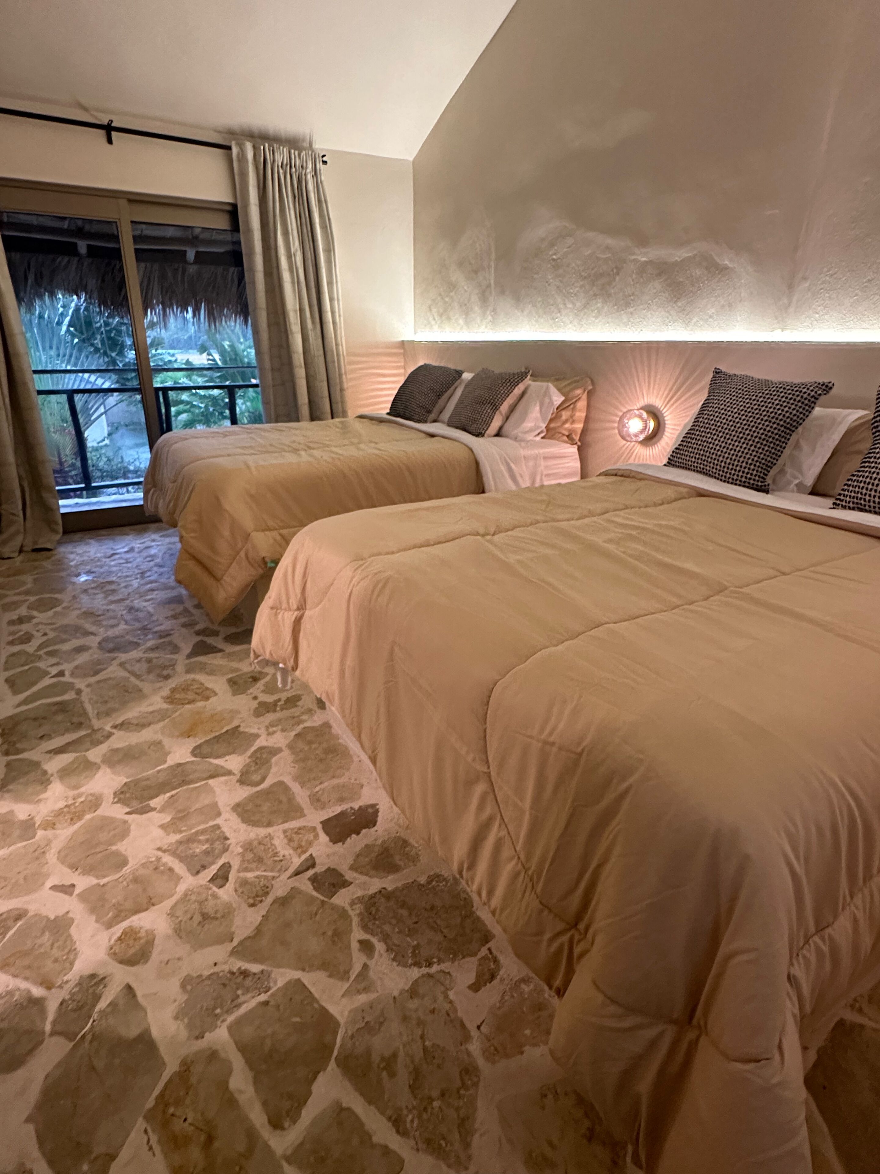 A bedroom with two beds, stone flooring, a large window with curtains, and a wall-mounted light fixture.