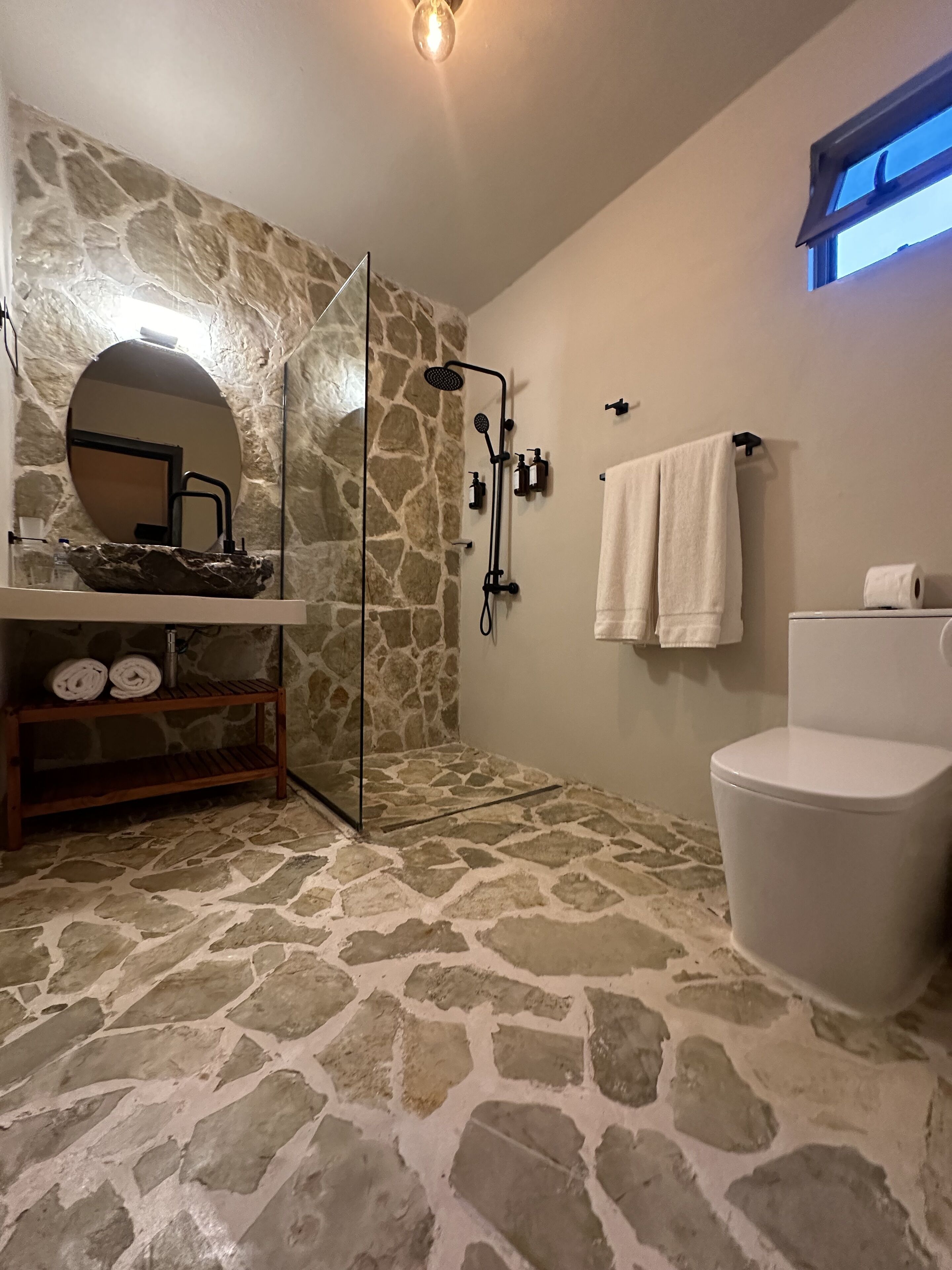 Uma casa de banho com chão de pedra, sanita branca, chuveiro com cabeça de duche manual, lavatório com espelho e toalhas penduradas numa barra.