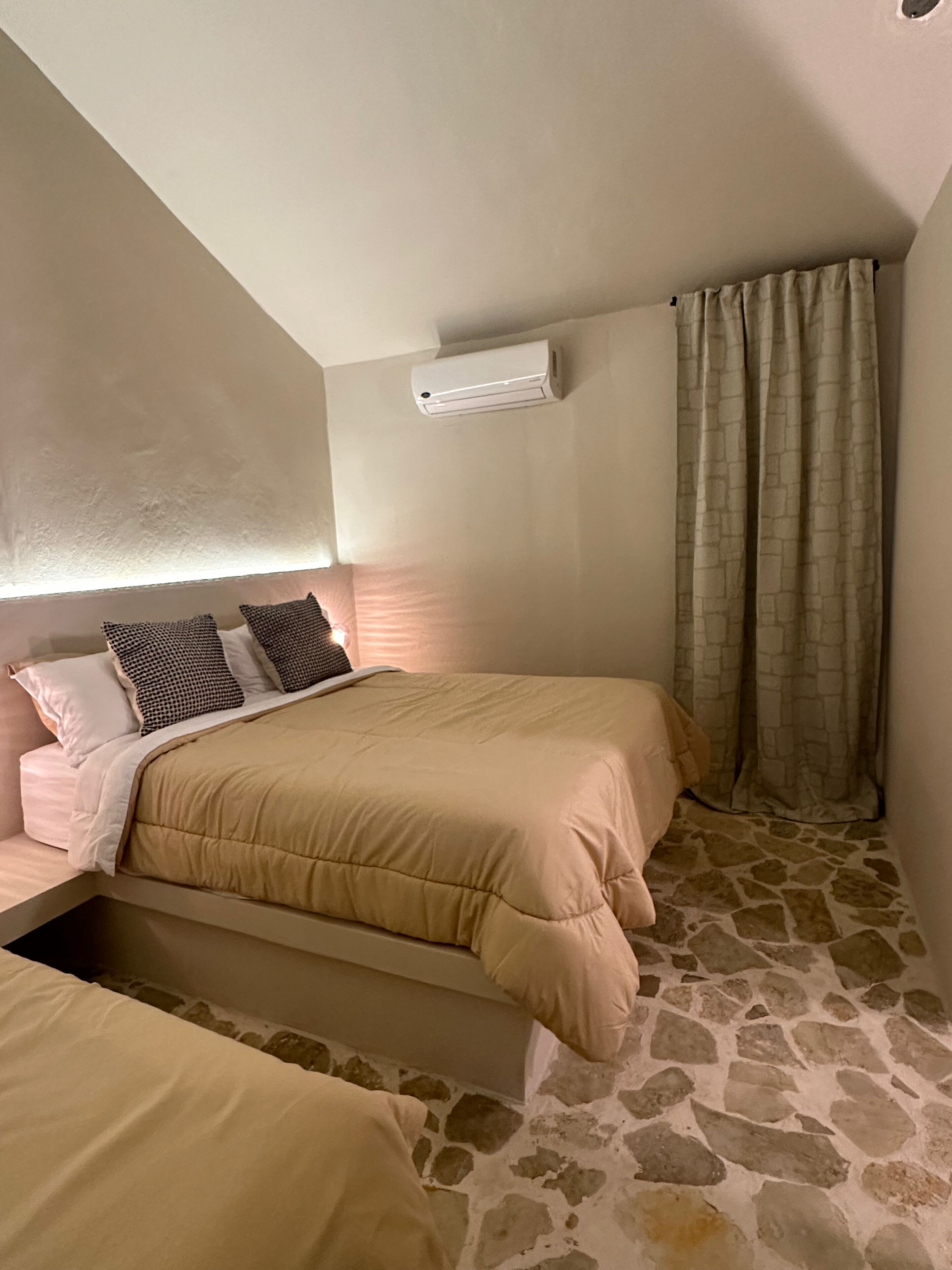 A bedroom with a bed, a small window with curtains, a wall-mounted air conditioner, and a stone-patterned floor.