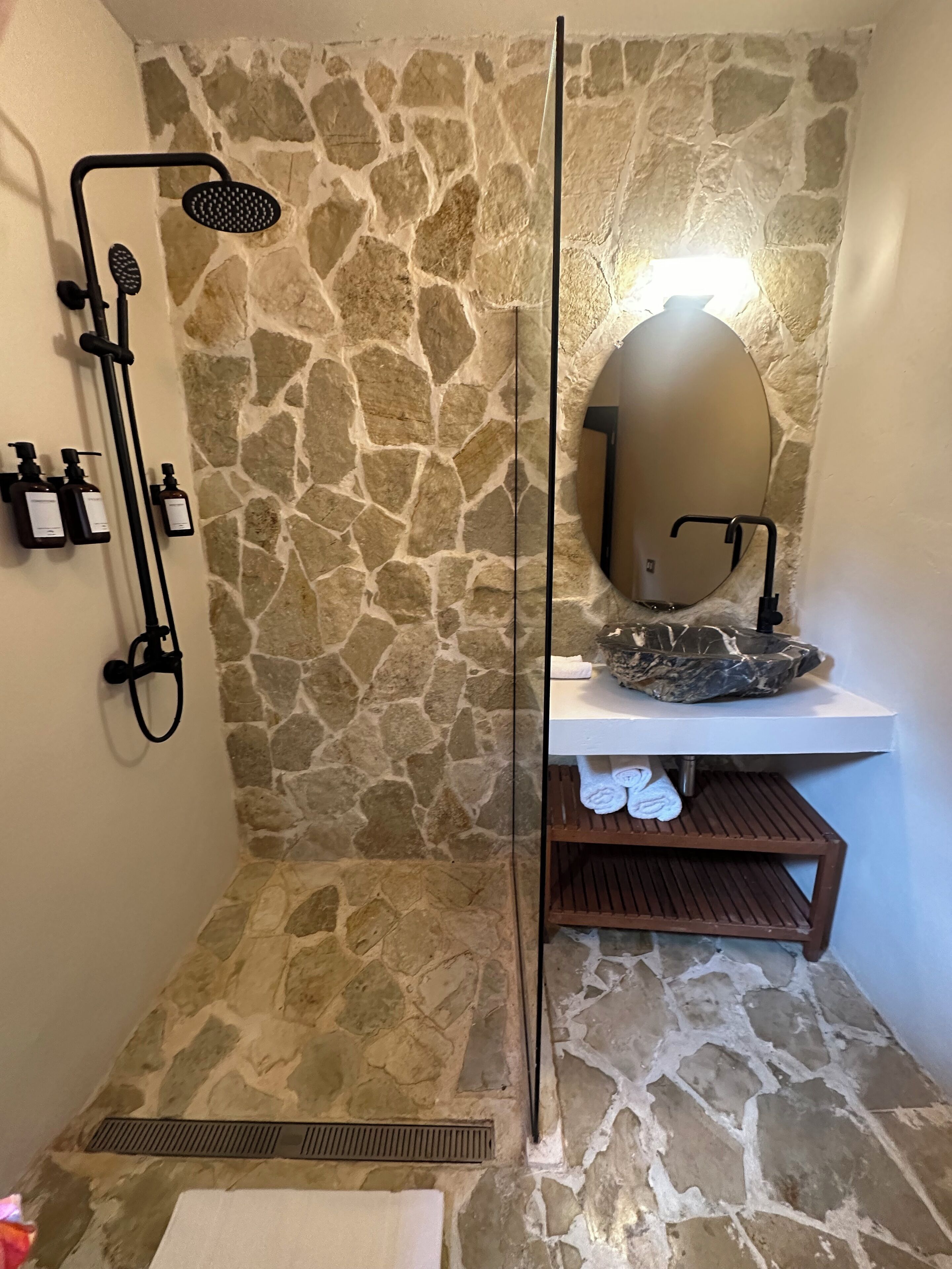 A stone-walled shower area with a handheld showerhead, a wall-mounted mirror, and a shelf with towels.