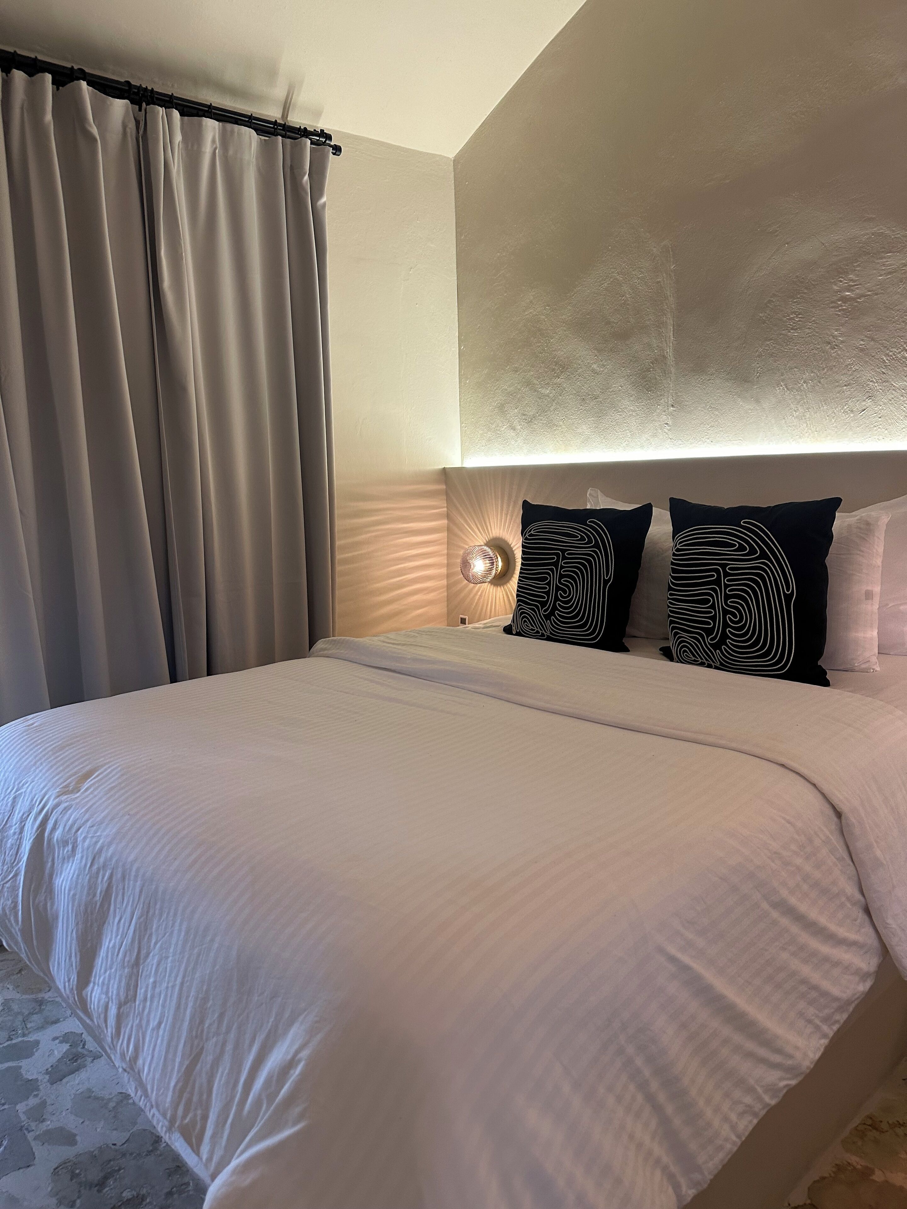 A hotel room with a large bed, a bedside lamp, and two black pillows with a unique design.