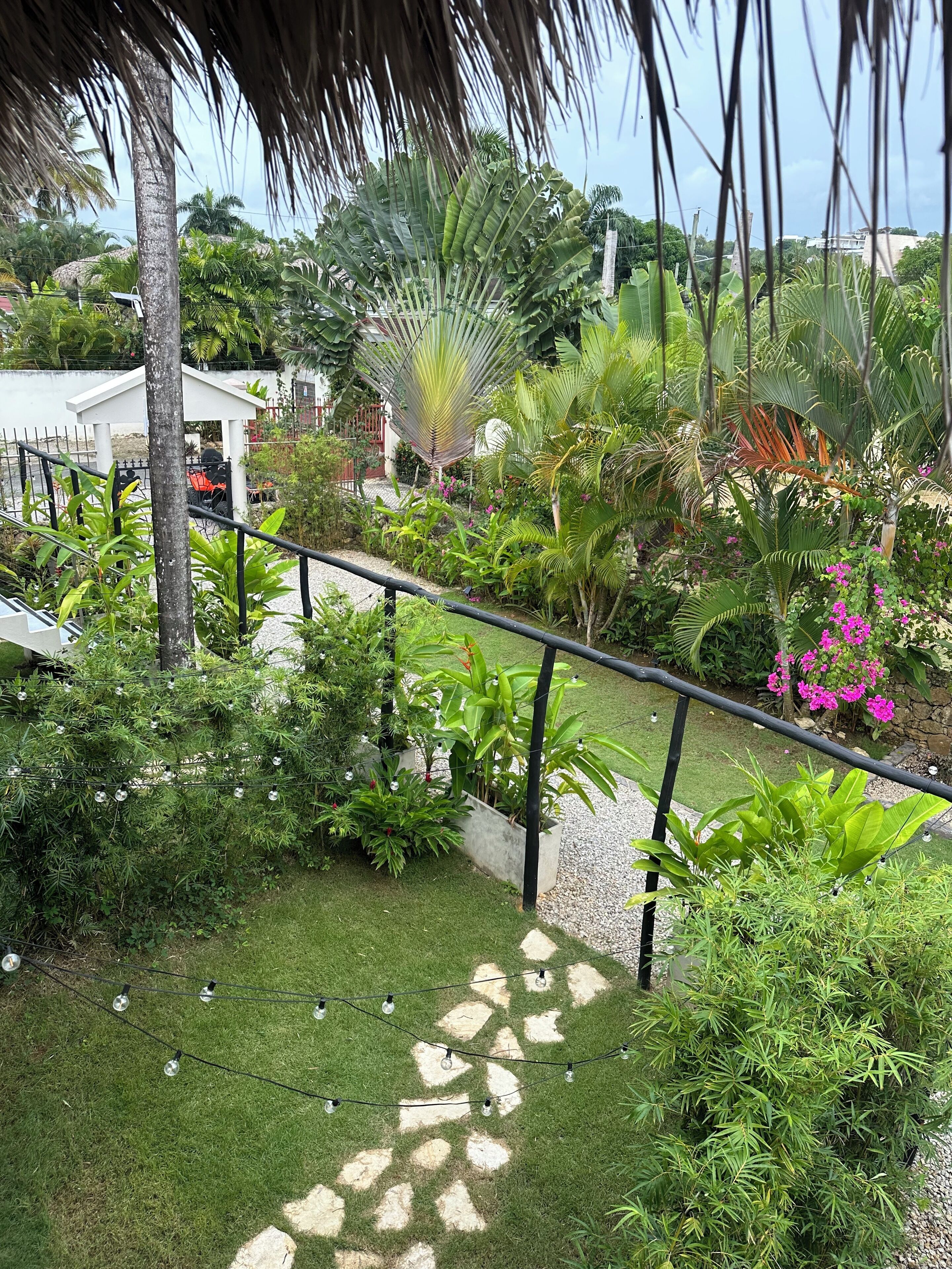 A lush garden with a variety of tropical plants and a hammock. There is a white gazebo and a thatched roof structure.