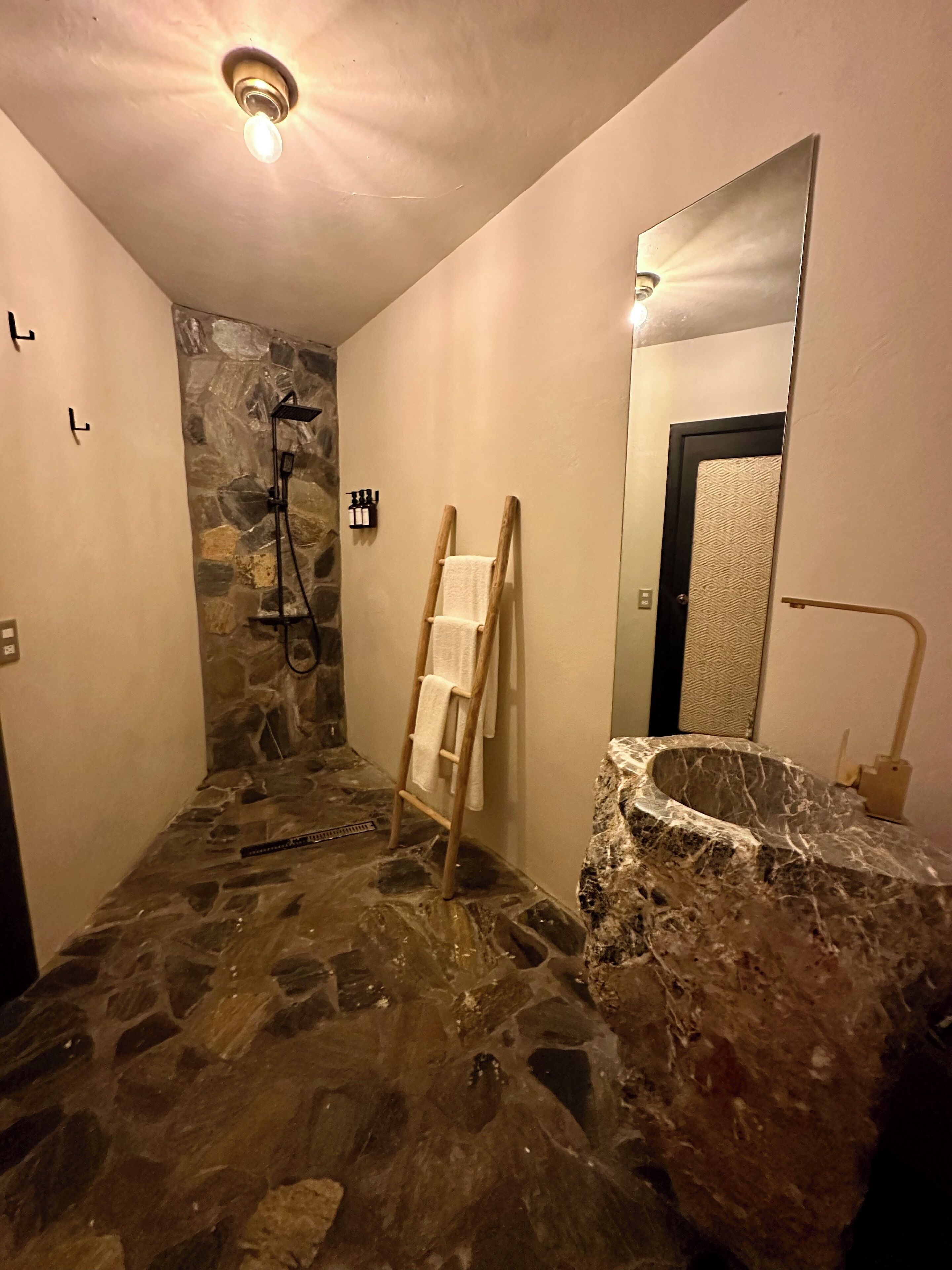 A bathroom with a stone floor, a wooden ladder, a towel rack, a shower, and a marble sink.