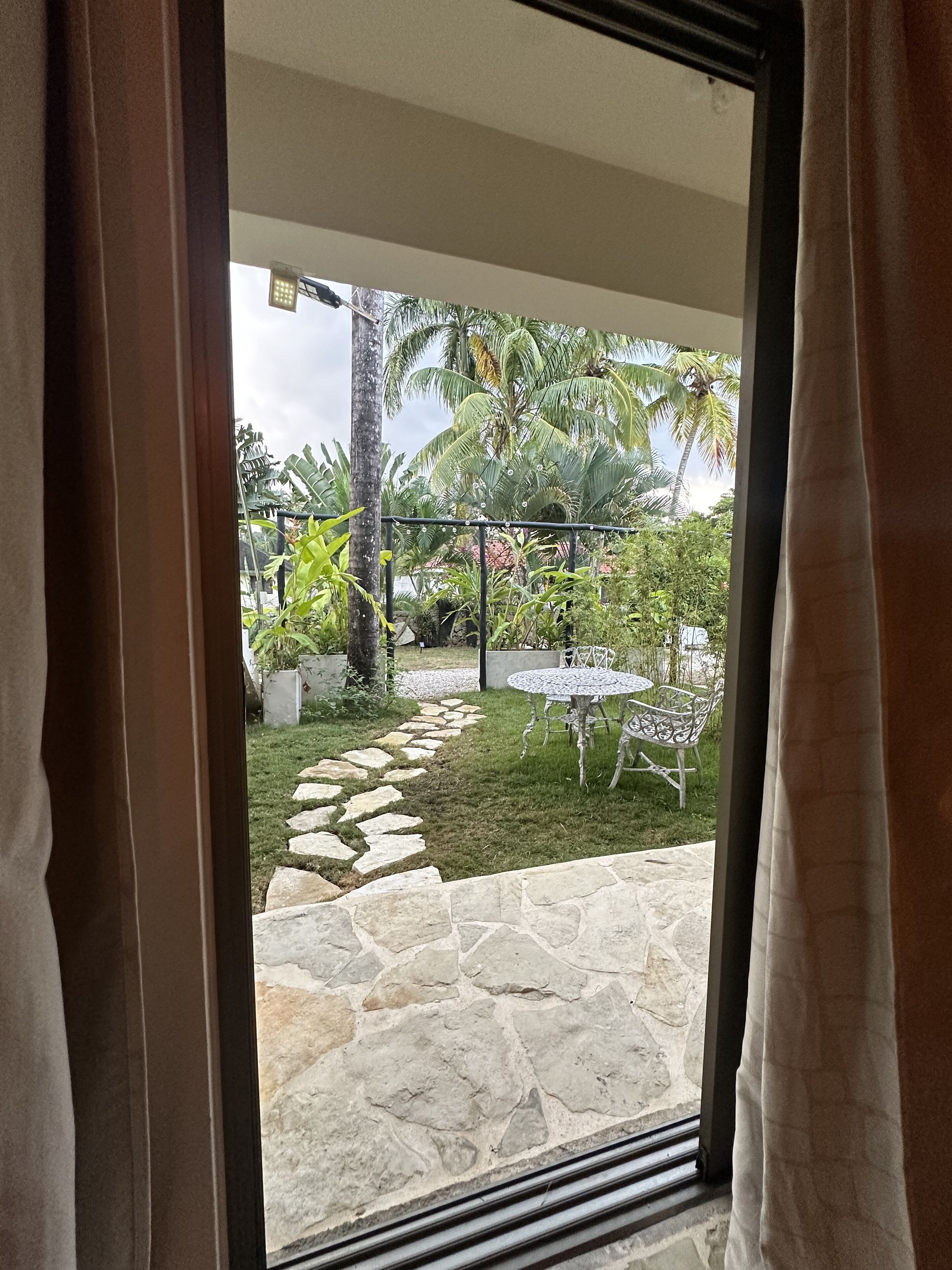 A view through a window of a stone patio leading to a grassy area with a table and chairs, surrounded by palm trees and a stone pathway.