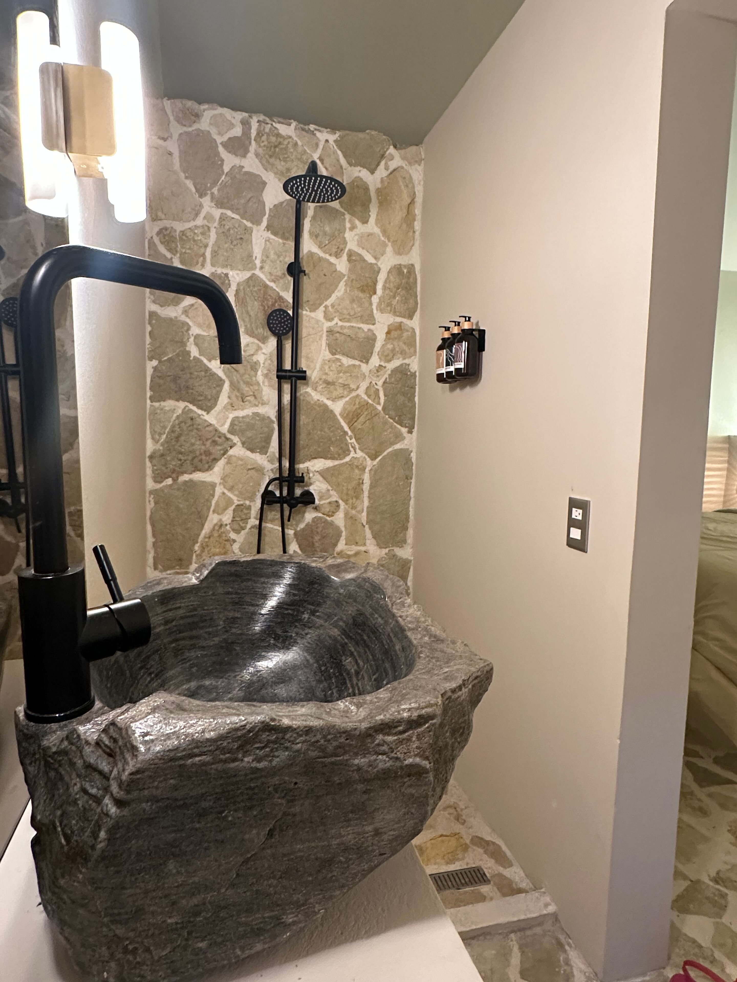 A stone sink with a black faucet and showerhead in a bathroom with stone walls and a white countertop.
