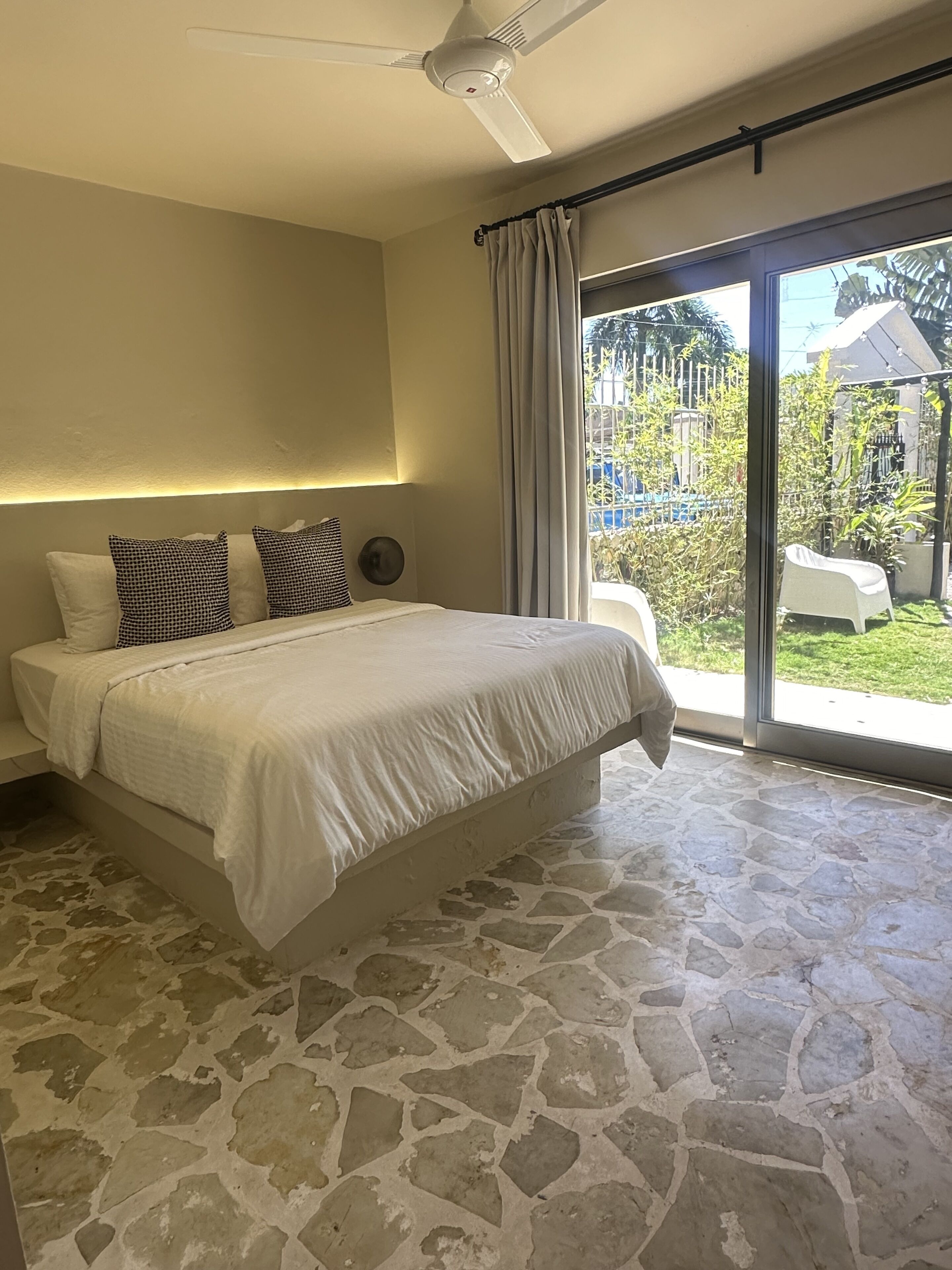 Ein Schlafzimmer mit Steinboden, einem Bett mit weißer Bettwäsche, einem Deckenventilator und Blick auf einen Garten durch eine Schiebetür.