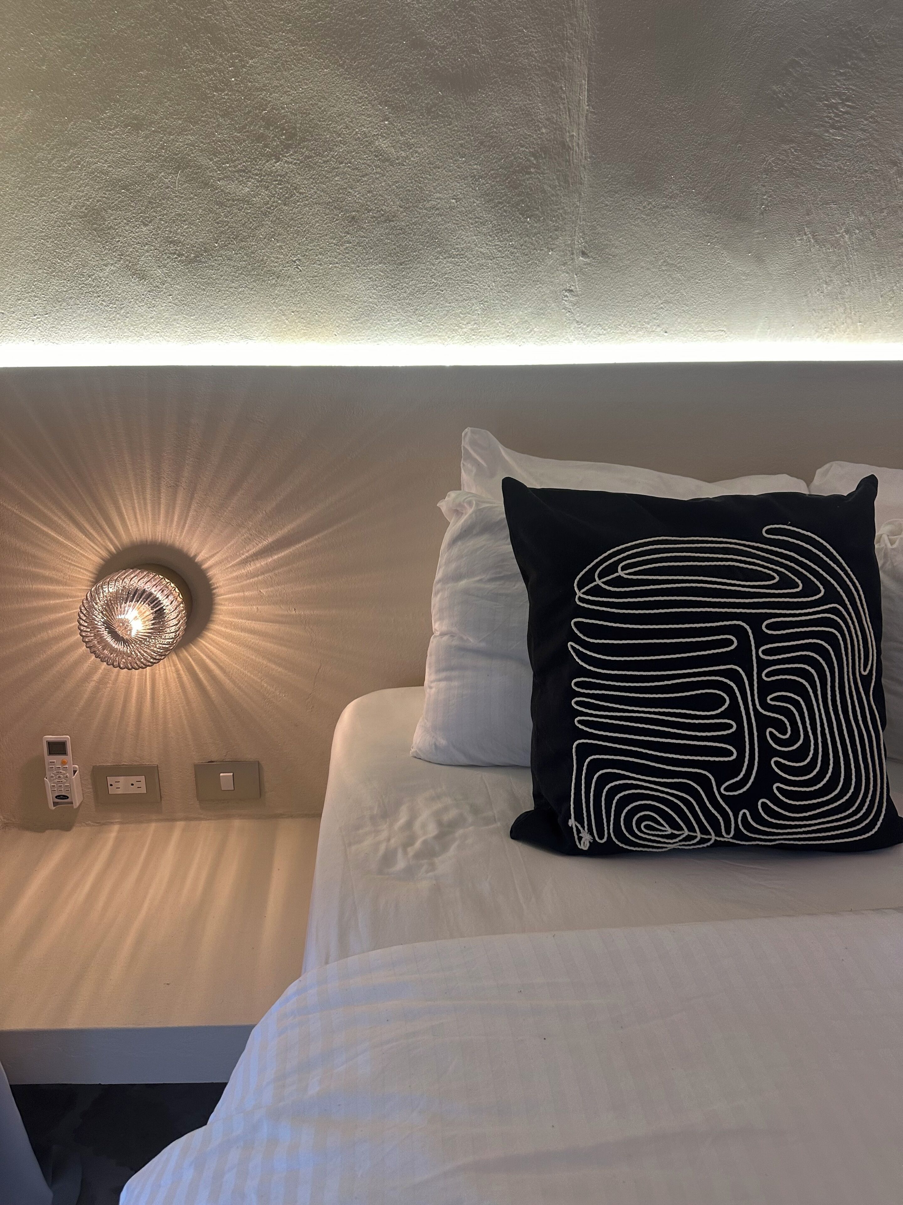 Un lit bien fait, avec un oreiller noir et blanc, un luminaire fixé au mur et un panneau de commande sur le mur.
