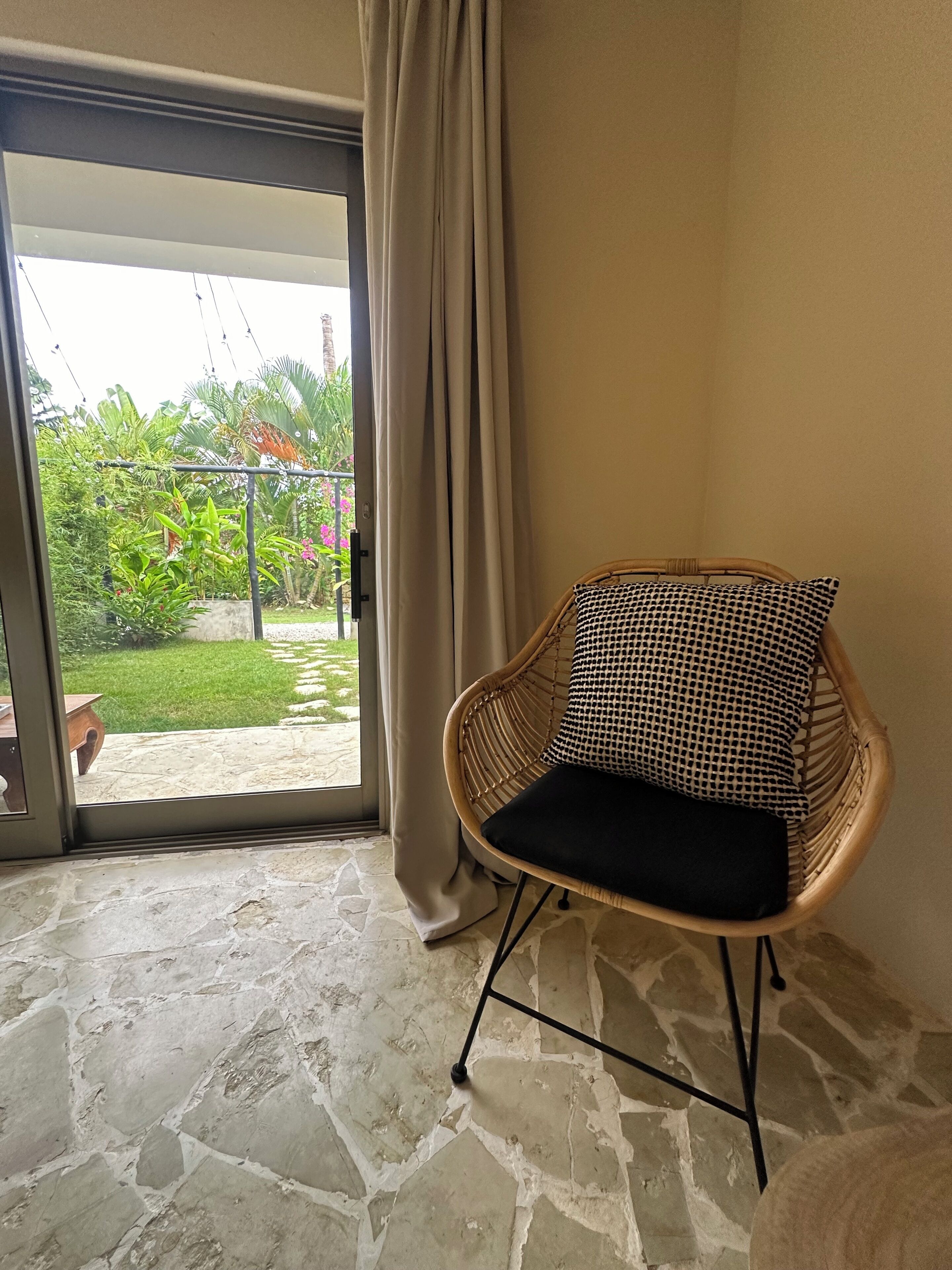 Une chaise en osier avec un coussin noir, dans une pièce dont le sol est recouvert de pierres, et qui offre une vue sur un jardin à travers une porte vitrée.