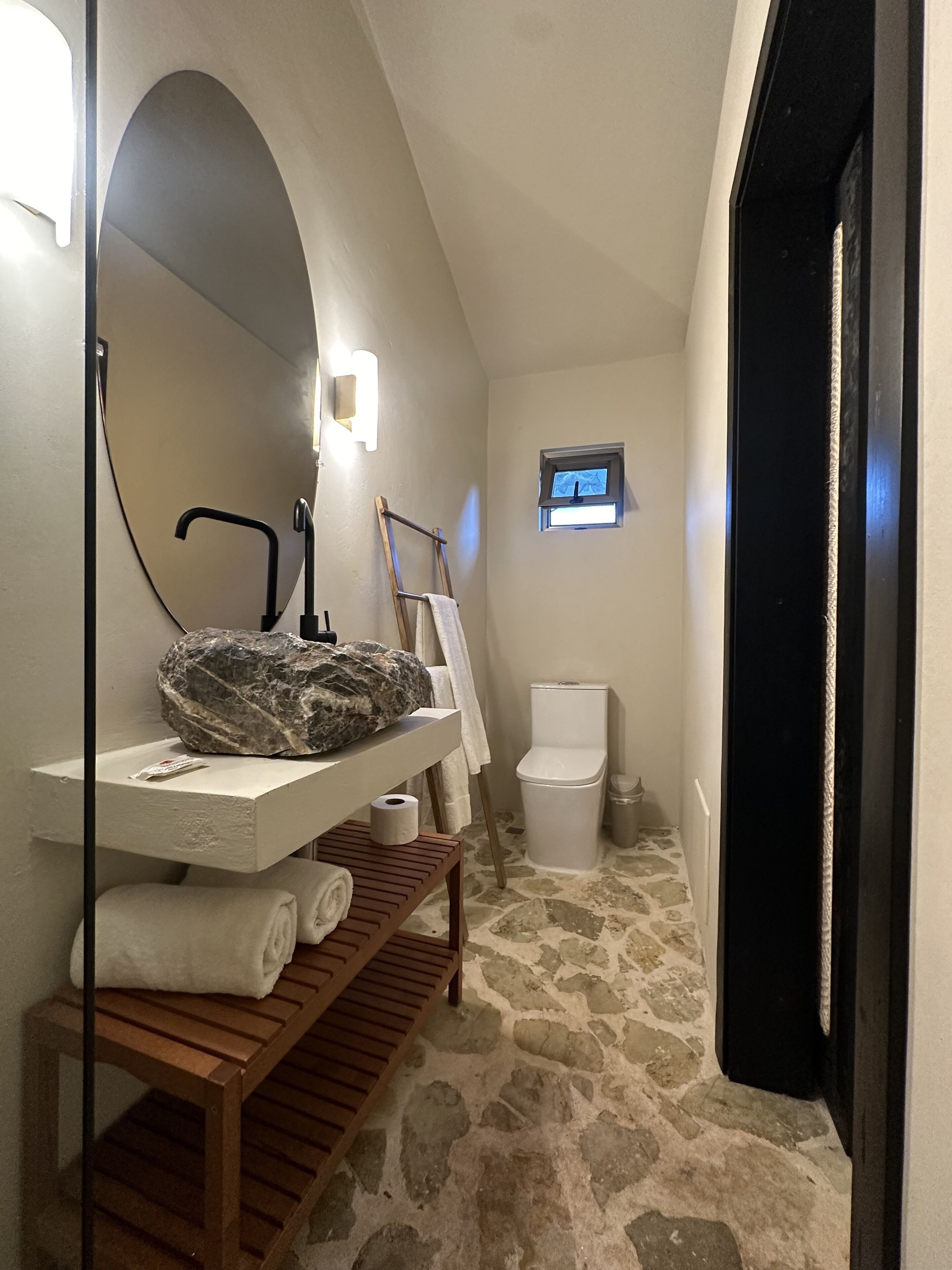 Un baño con suelo de piedra, un lavamanos blanco con encimera de mármol, un banco de madera, un perchero, un inodoro y una pequeña ventana con un televisor.